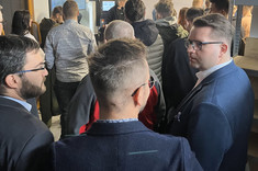 Grupa uczestników w hali produkcyjnej, niektórzy wymieniają uwagi, inni obserwują prezentację na monitorze,